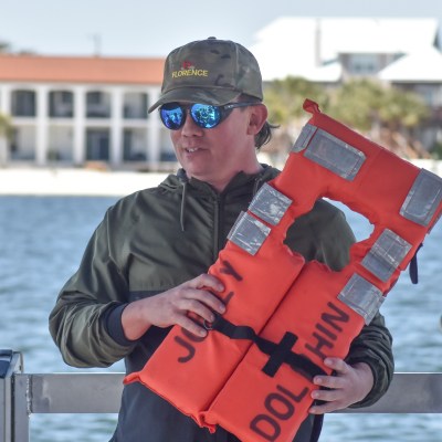 Person holding an orange life jacket on a boat with buildings and water in the background.