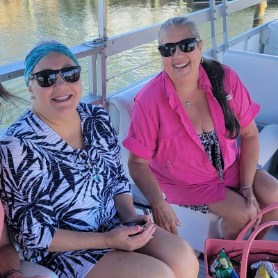 Three women sitting on a boat, smiling and wearing sunglasses, with bags and a cooler nearby.
