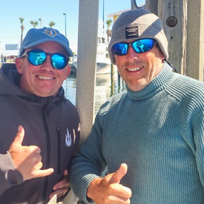 Two men in sunglasses and hats smiling at a dock with boats in the background.