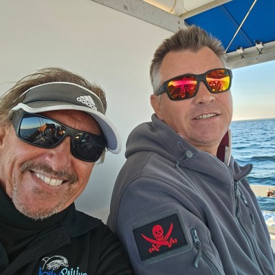 Two men smiling on a boat, wearing sunglasses. One has an Adidas visor, the other wears a jacket with a skull patch.