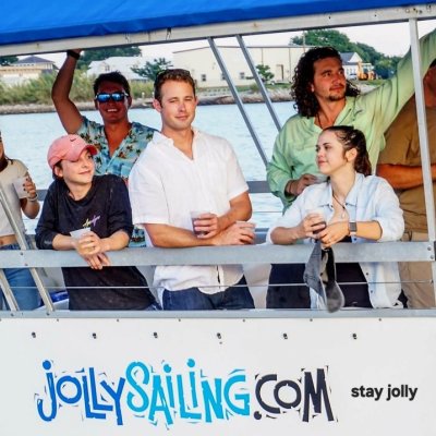 Group of people on a boat with a 'JollySailing.com' sign, smiling and holding drinks.