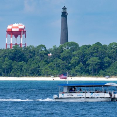 Boat on water with lighthouse and red-striped towers in background.