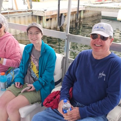 Three people sitting on a boat bench, smiling, with water and a dock in the background.