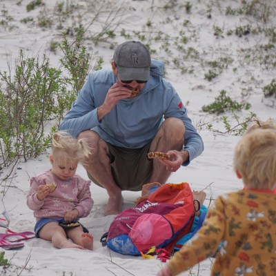 Three people sitting on a beach with snacks and bags around them.