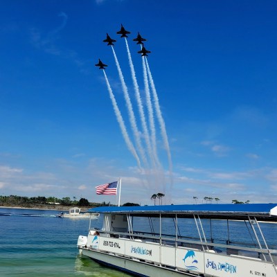 Five jets in formation emit trails above a boat with an American flag on a sunny day.