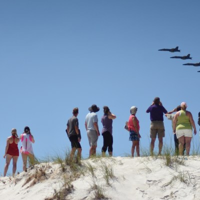 People on a sandy dune watching four jets flying in formation in a clear blue sky.