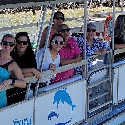 Group of people smiling on a boat with canopy, near rocky shore.