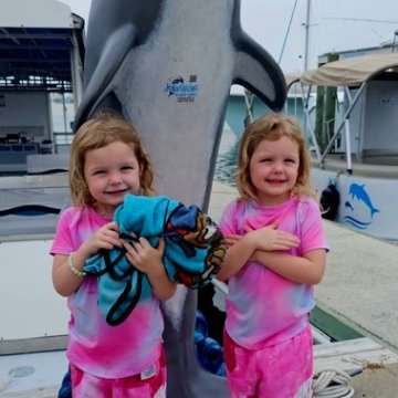 Two smiling kids in matching pink outfits stand by a dolphin statue near a marina.
