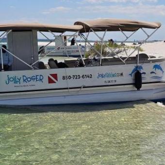 Boat named 'Jolly Roger' anchored on sandy beach with people nearby, clear water and sunny sky.