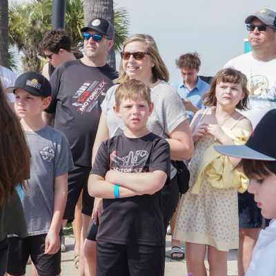 Group of people outdoors, some wearing baseball caps and sunglasses, with palm trees and a blue building in the background.