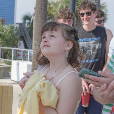 Girl in sundress holding a towel, surrounded by people outdoors.