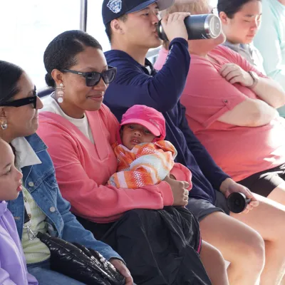 Group of people sitting on a boat, including women, children, and a man drinking from a bottle.