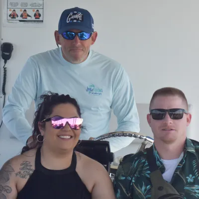 Three people on a boat, two seated, all wearing sunglasses and casual attire.