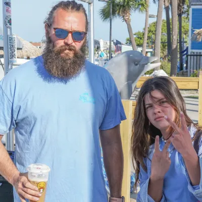 Man with a beard and sunglasses holds a drink; woman next to him flashes peace signs in a sunny outdoor setting.