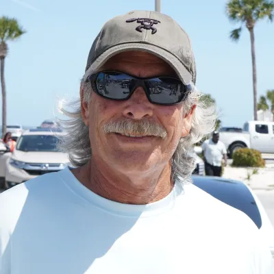 Man with a mustache and sunglasses smiles outdoors on a sunny day, palm trees and cars in the background.