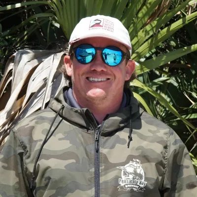 Person in camouflage jacket, sunglasses, and cap standing in front of palm leaves.