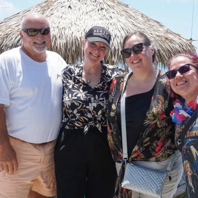 Four people smiling on a dock beside a thatched-roof hut and boats.