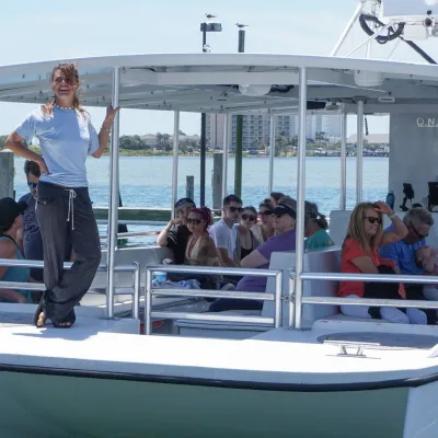 People sitting on a boat in a marina, with a smiling woman standing at the front.