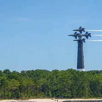 Four jets in formation flying above trees with white vapor trails.