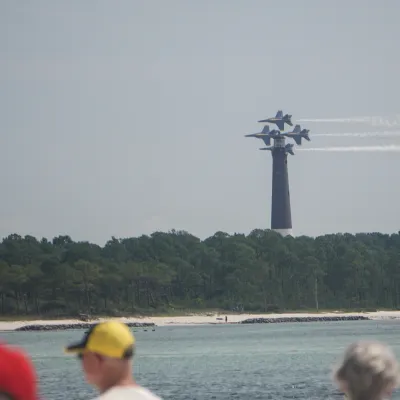 Three jets flying in formation over water and forest with spectators in foreground.