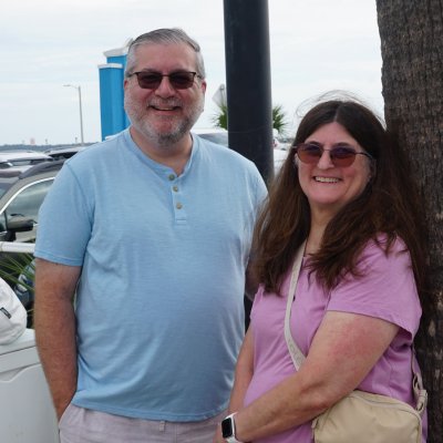 Two people smiling near a palm tree by parked cars.