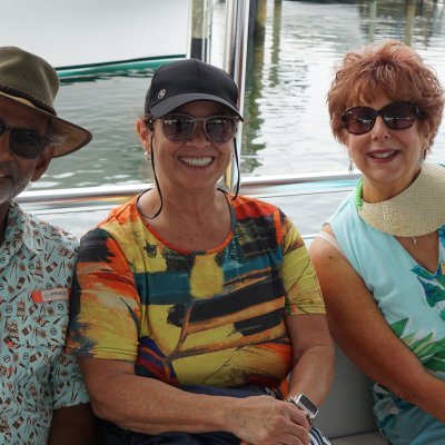 Three smiling people sitting on a boat, wearing casual summer clothes and sunglasses.