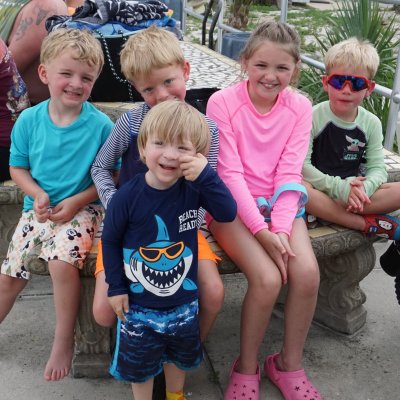 Five smiling children sit on a stone bench outdoors, wearing colorful casual clothes.