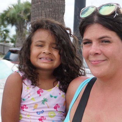 Smiling woman holding a child outdoors near palm trees and parked cars.