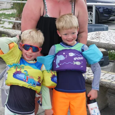 Two kids in life jackets and swimwear, smiling outdoors with an adult behind them.