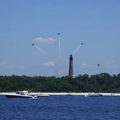 Jets in formation above a lighthouse with boats on the water below.