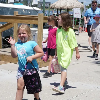 Children walking on a dock, some holding bags, with boats in the background.