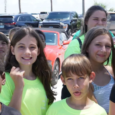 Group of children and adults standing in front of parked cars on a sunny day.