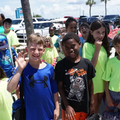 Group of children smiling outdoors in bright shirts near palm trees.