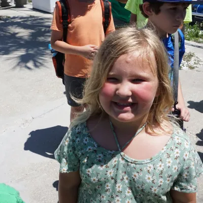 Smiling child outdoors with group, wearing a green floral shirt.