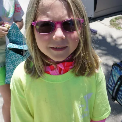 Young girl with pink sunglasses and neon shirt standing outdoors.