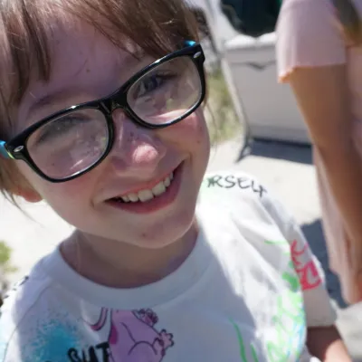 Child with glasses smiling outdoors, wearing a graphic shirt.