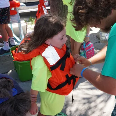 Adult helps child put on an orange life jacket outdoors.