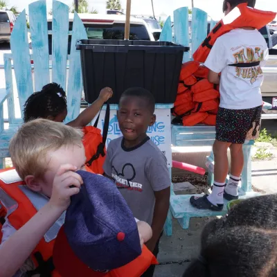 Children wearing life jackets near large chair on a sunny day, with cars and buildings in the background.