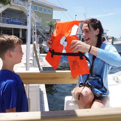 Person giving life jacket to child on a dock beside a boat.