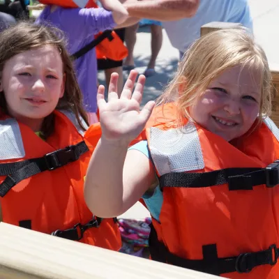 Two children in orange life jackets, one waves, standing by a wooden railing.