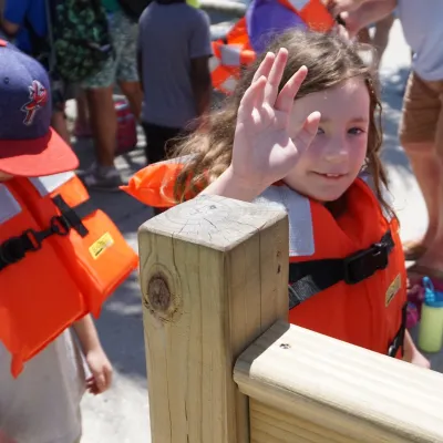 Children in orange life vests, one waving, near a wooden railing.