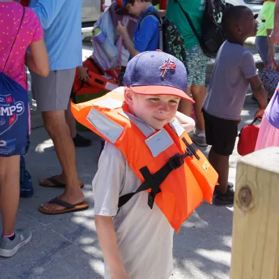 Child in life jacket and cap with group of people outdoors.